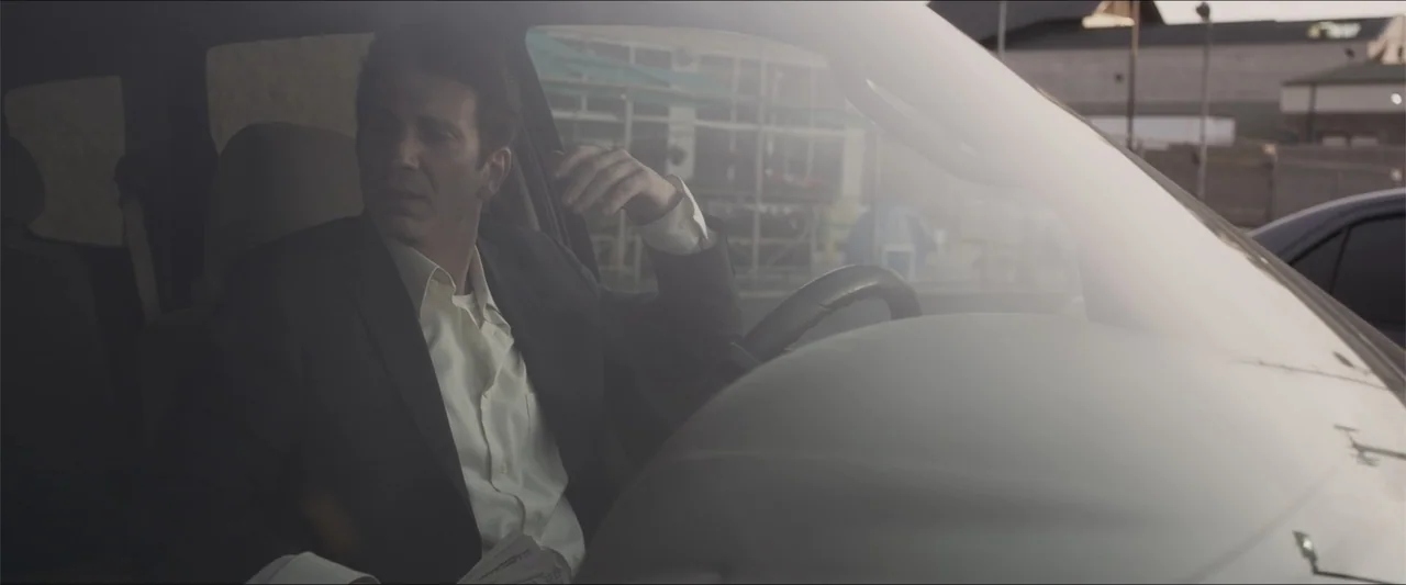 Heavenly Deposit (2019) movie still: medium — The interior of a car during twilight with a blurred building in the background. A man in a suit sits…