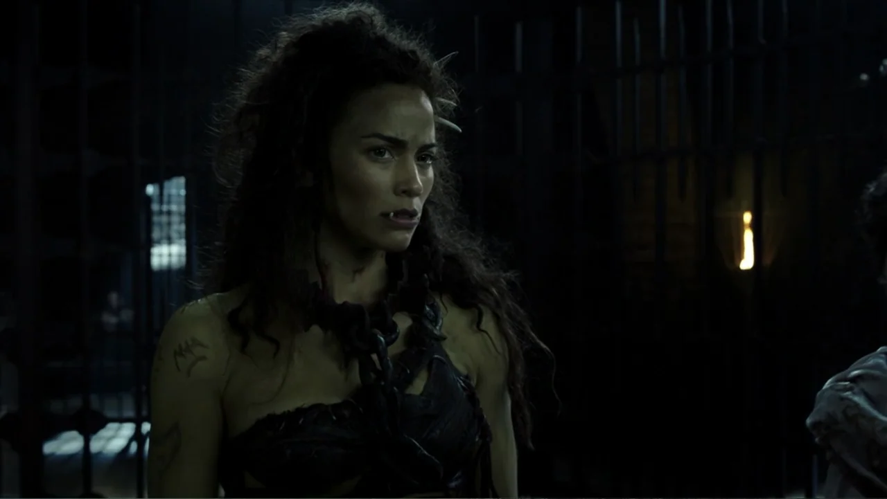 Warcraft (2016) movie still: medium — A dark dungeon setting with heavy iron barred gates visible in the background. A woman wearing a leather…