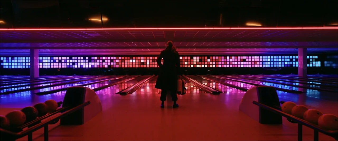 Gunpowder Milkshake (2021) movie still: fullbody — An empty bowling alley features lanes illuminated by intense red and purple neon lights. A woman…
