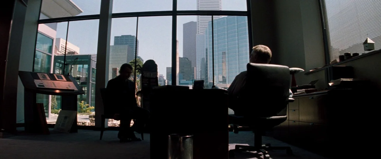 In The Mouth of Madness (1994) movie still: wide — A spacious high-rise office features floor-to-ceiling windows overlooking a city skyline. Two men…