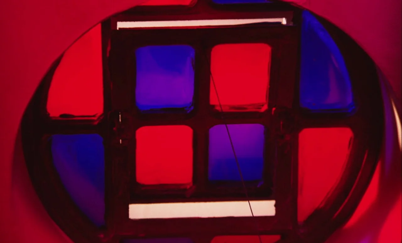 Mahler (1974) movie still: closeup — A close-up view shows a grid of red and blue translucent panels. The window frame is surrounded by an oval…