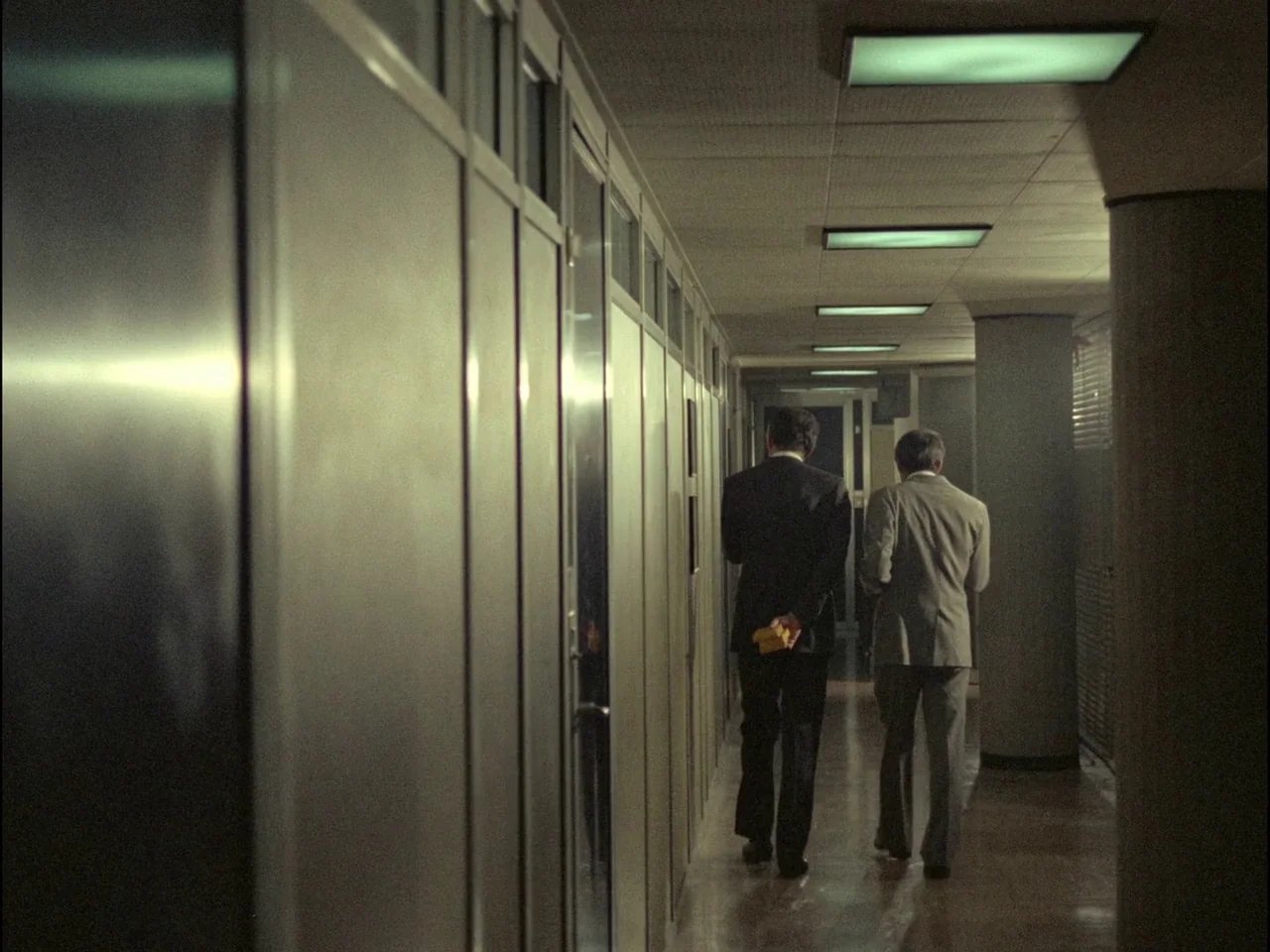 Dillinger is Dead (1969) movie still: fullbody — A narrow office corridor features metal-paneled walls and a repeating row of ceiling lights. Two…