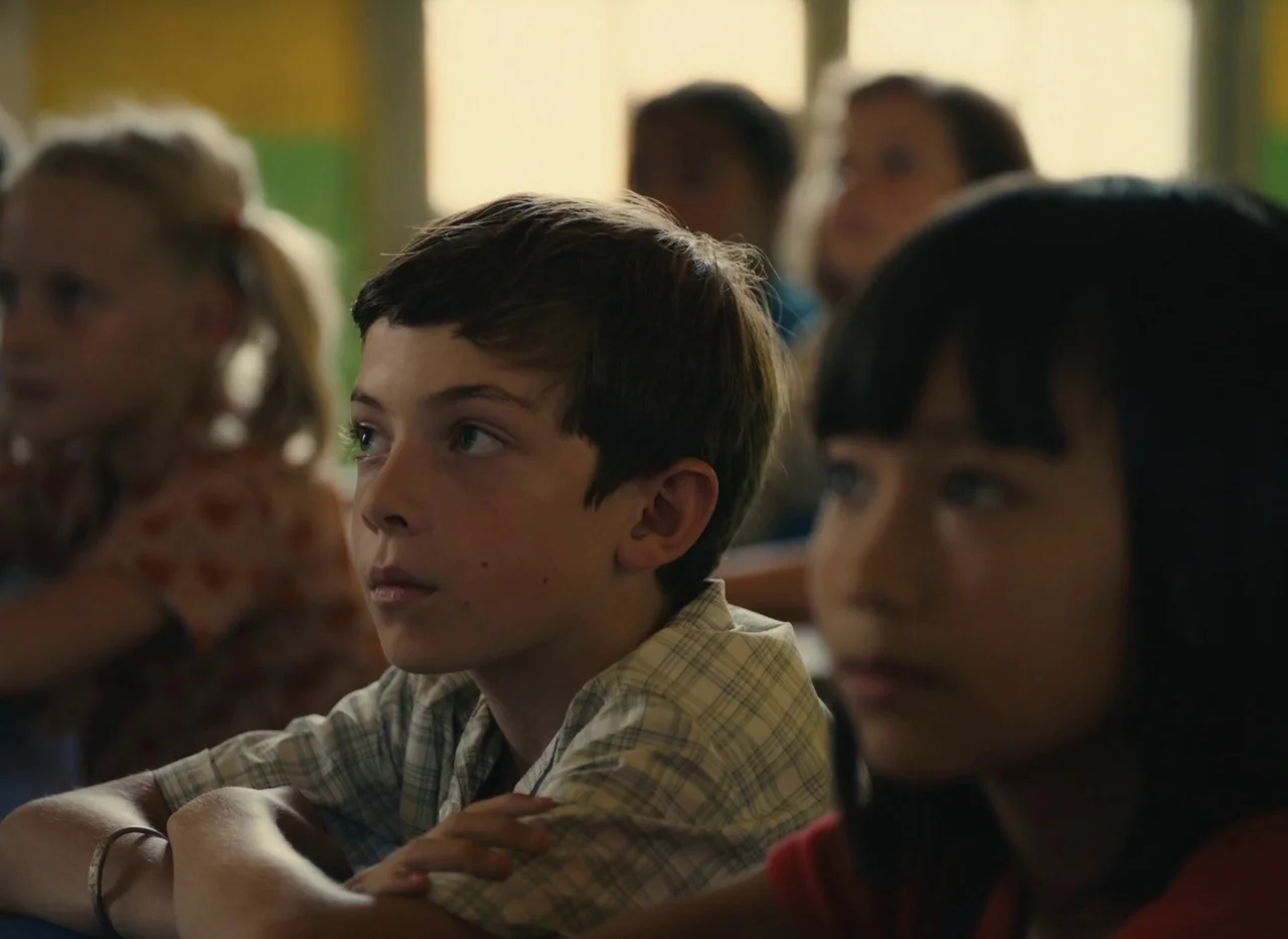 Red Island (2023) movie still: medium — A classroom interior with several children sitting at desks in a row. A boy in the foreground looks forward…