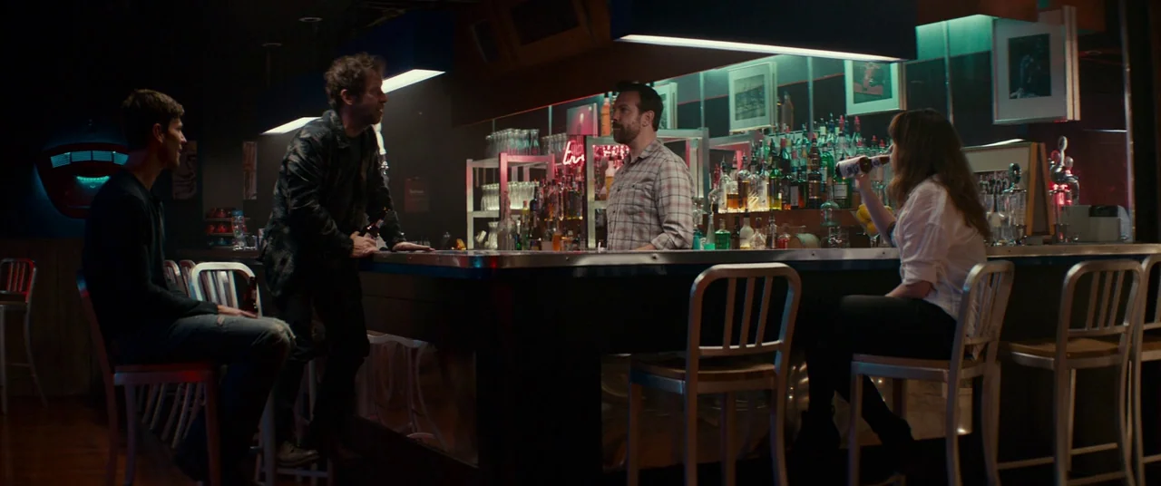 Colossal (2016) movie still: wide — A dimly lit bar counter is filled with various glass bottles and barware supplies. Four people are positioned…