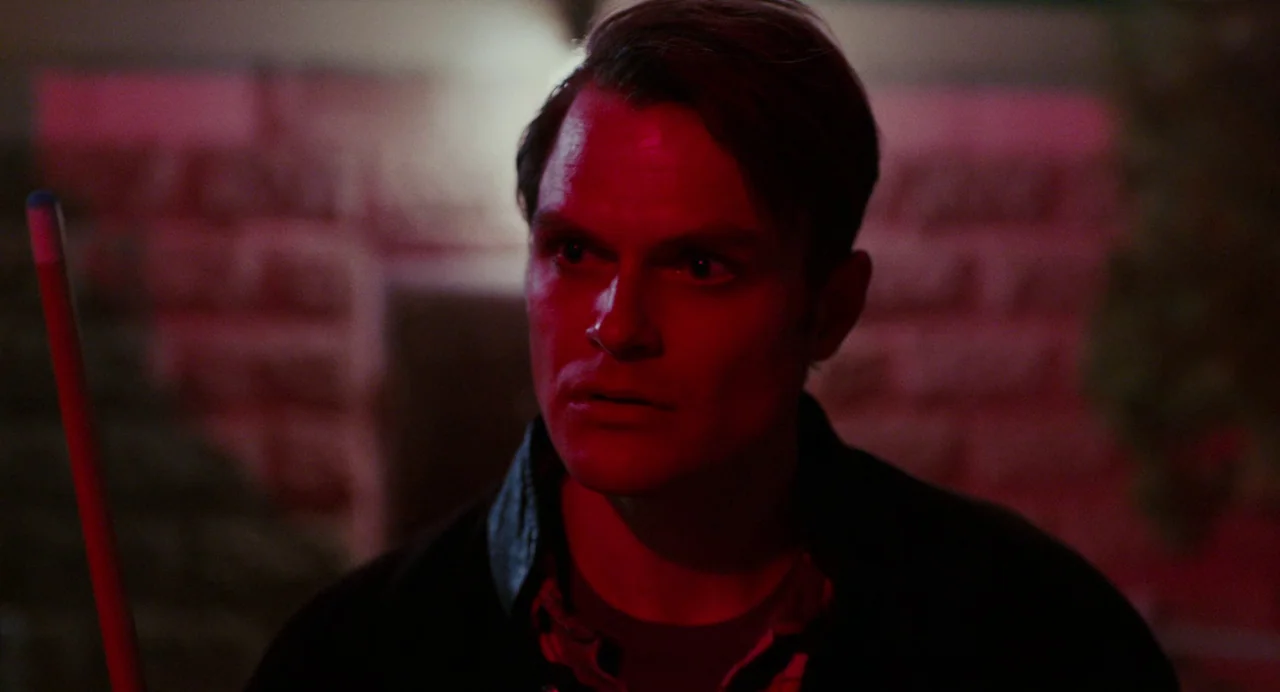 My Animal (2023) movie still: closeup — A dimly lit space features a stone wall texture washed in saturated red light. A man holds a pool cue and…
