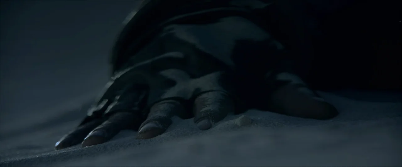 Monster Hunter (2020) movie still: closeup — A dark, textured expanse of wind-swept sand covers the foreground and background of the frame. A gloved…