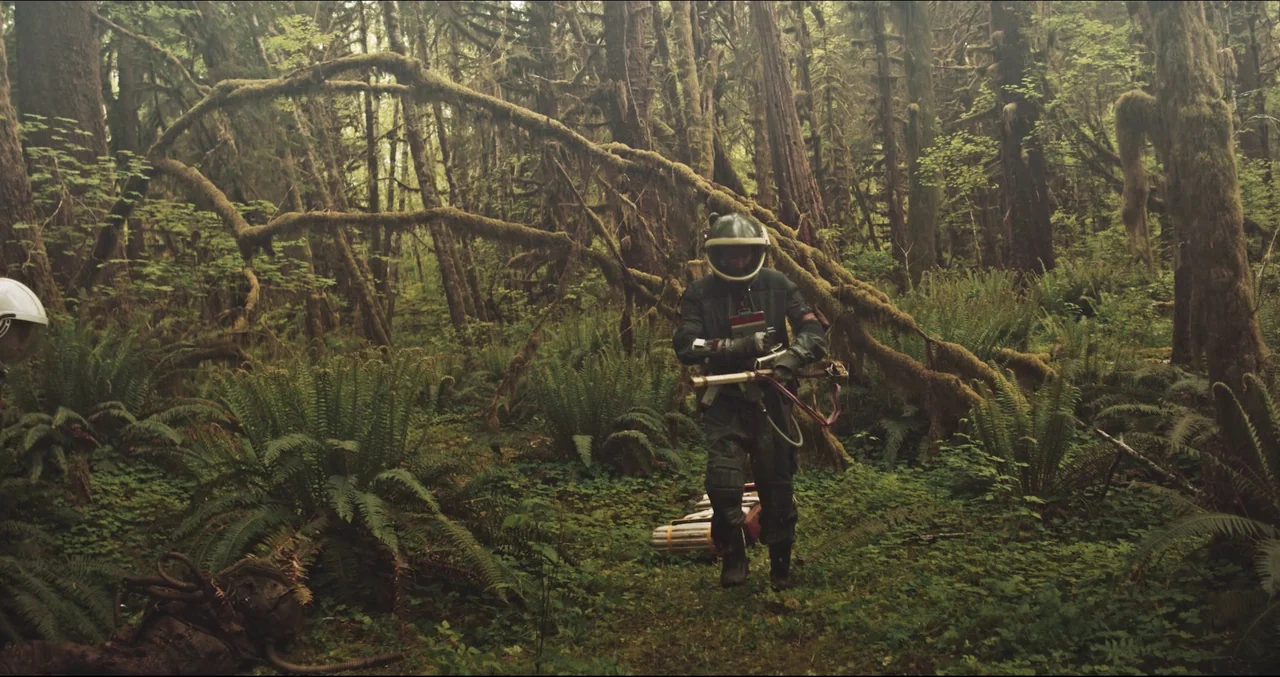 Prospect (2018) movie still: fullbody — A dense temperate rainforest features large ancient trees covered in thick, hanging moss. A person wearing a…