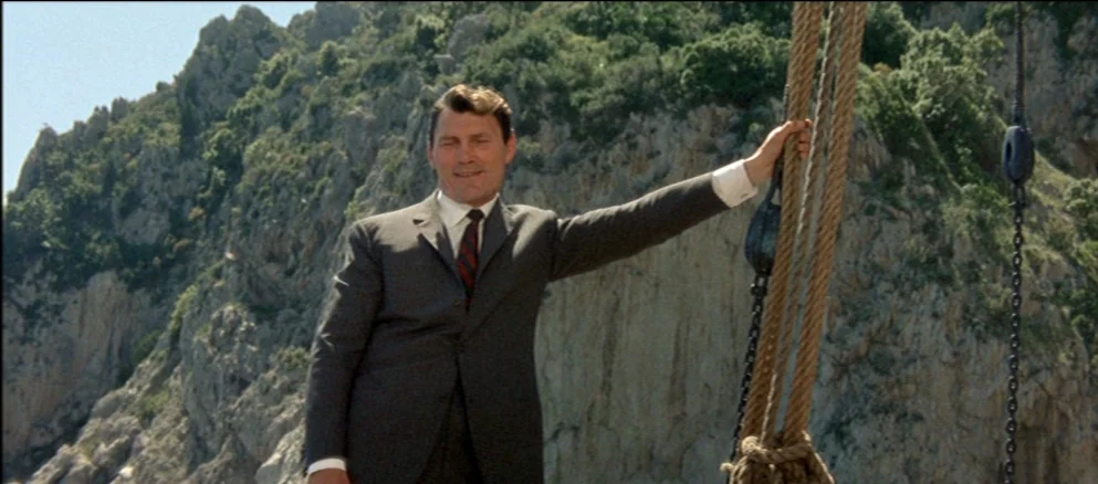 Contempt (Le Mépris) (1963) movie still: medium — A man in a business suit stands on the deck of a boat. Behind him rises a tall rocky cliff face…