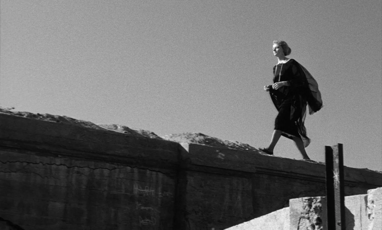 Night Tide (1961) movie still: fullbody — A woman in a black dress walks along the top of a stone ledge. The structure is set against a clear,…