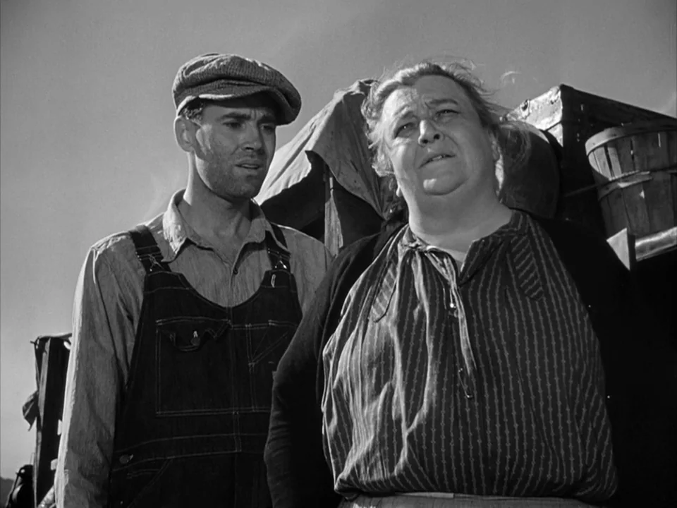 The Grapes of Wrath (1940) movie still: medium — Two individuals stand in an outdoor setting with wooden farm equipment behind them. The man wears…