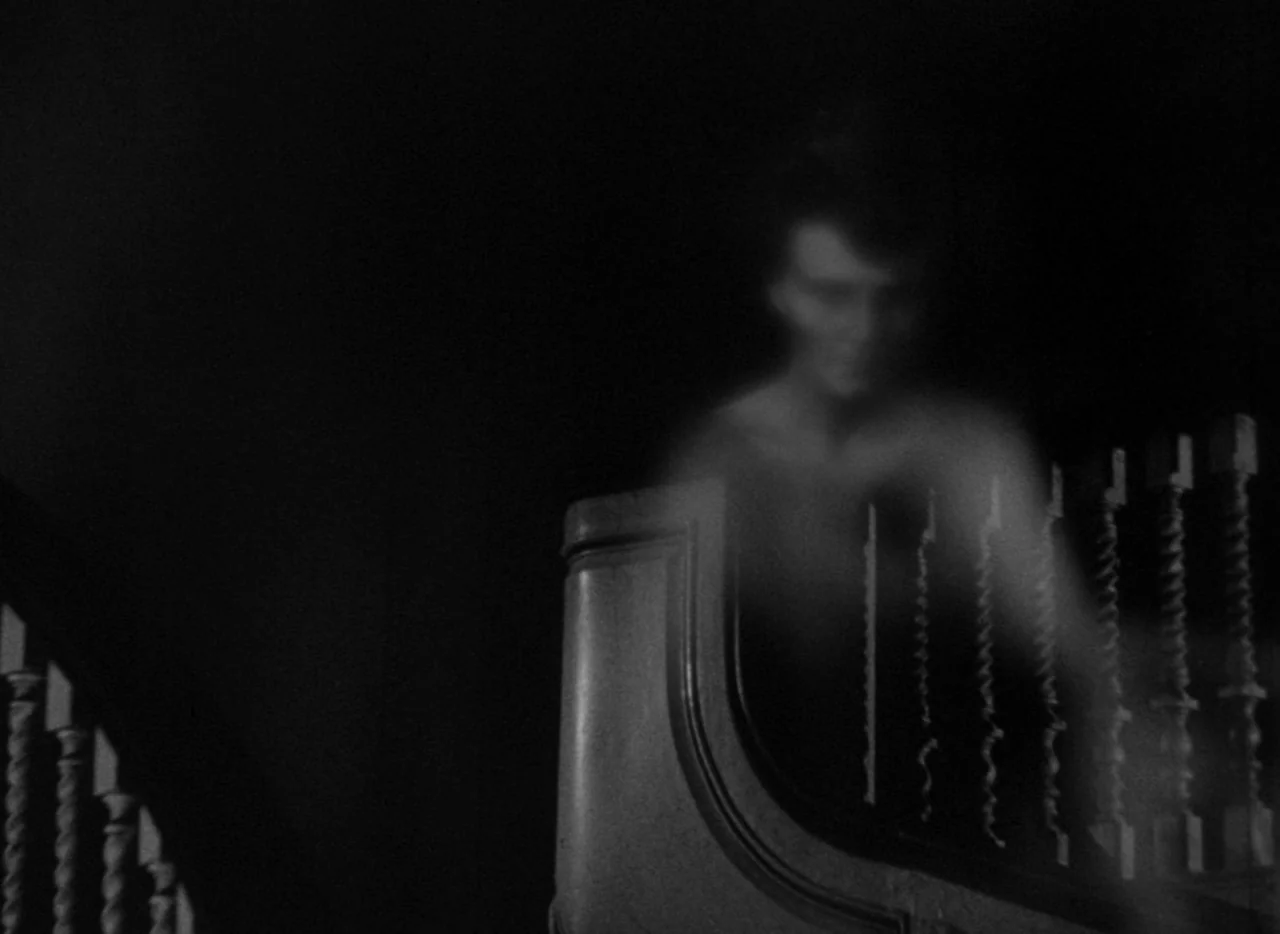 The Uninvited (1944) movie still: medium — A dark staircase interior features wooden banisters and a curved rail in the foreground. A ghostly,…