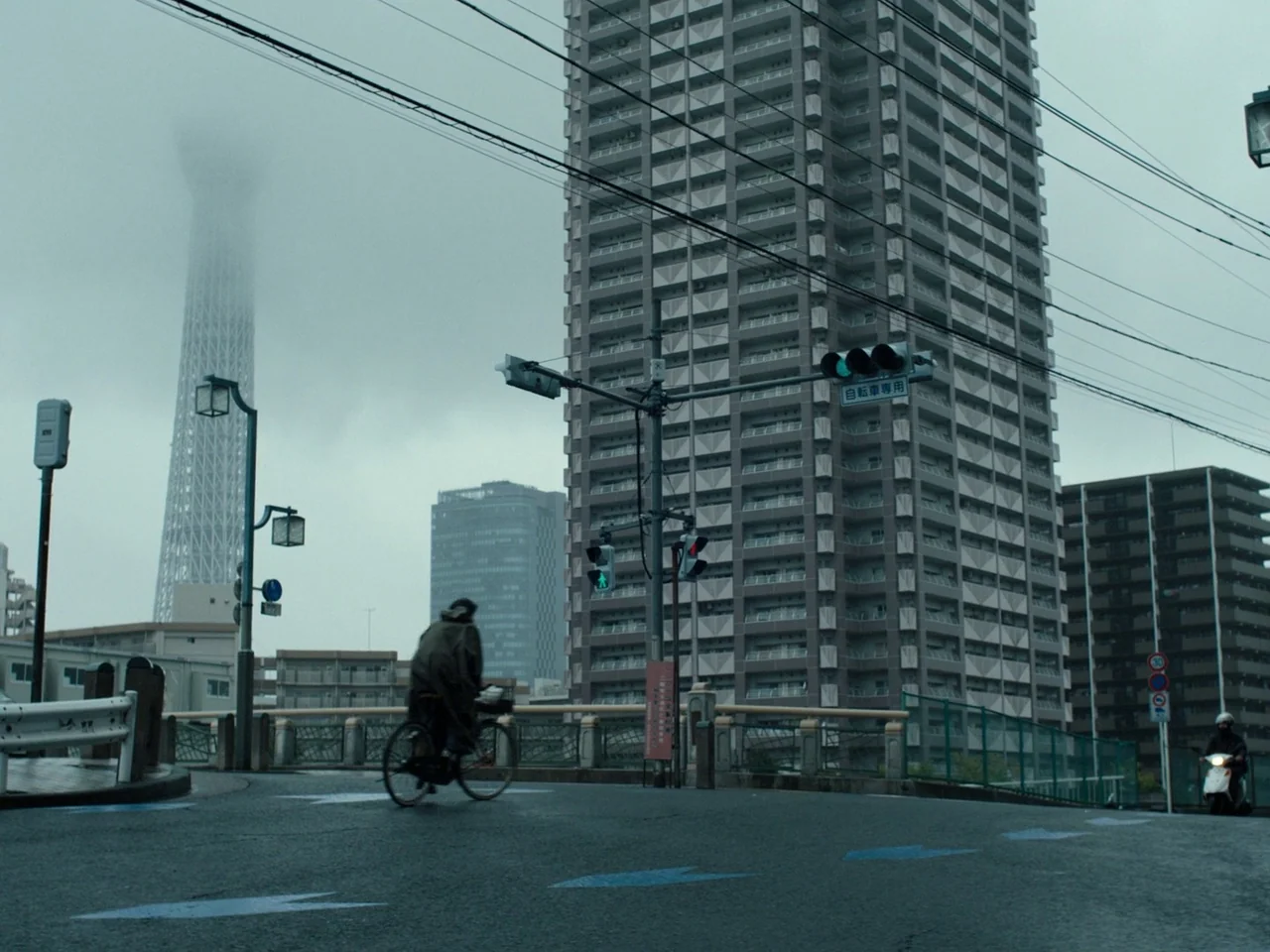 Perfect Days (2023) movie still: wide — A wet asphalt urban intersection features a tall residential tower and a foggy skyline. A cyclist travels…