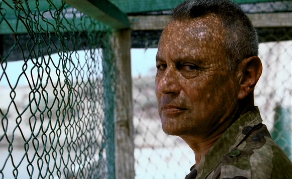 Beau Travail (1999) movie still: closeup — An outdoor area with a green chain-link fence and wooden support posts. An elderly man in military-style…