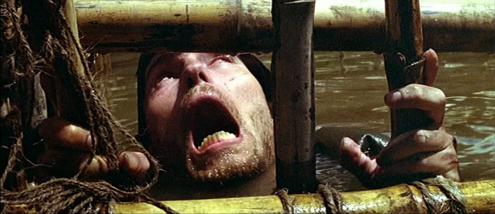 The Deer Hunter (1978) movie still: closeup — A man is partially submerged in murky water behind a bamboo cage structure. The subject is screaming…