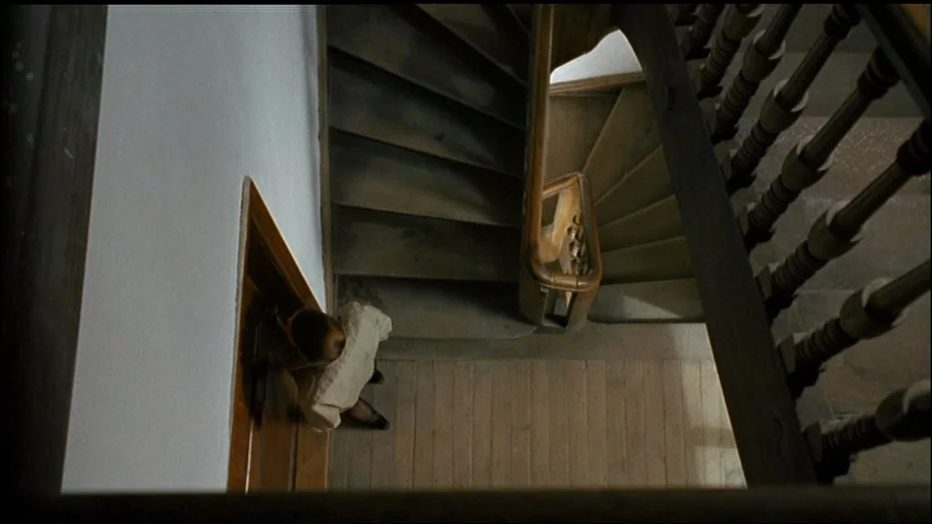 Chinese Roulette (1976) movie still: wide — A top-down view of a vintage wooden stairwell with a dark wooden banister. A woman in a blouse stands on…