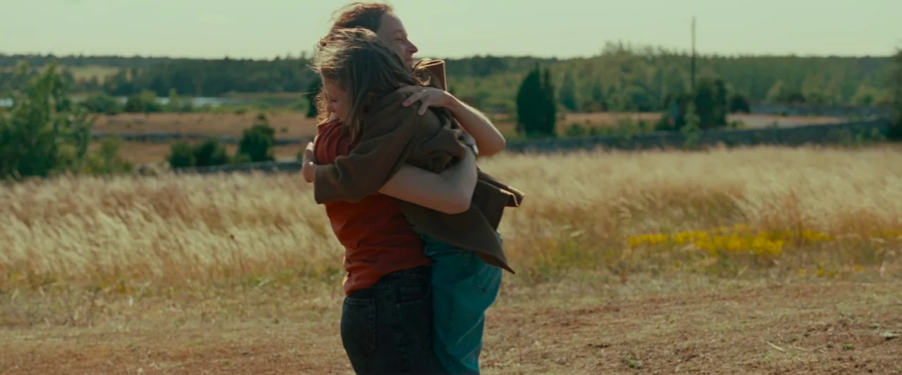 Bergman Island (2021) movie still: medium — A dry, grassy field extends toward a distant treeline under a bright, clear sky. Two people stand in the…