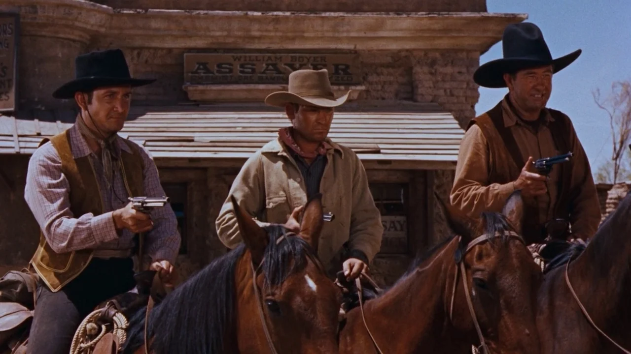 Rio Bravo (1959) movie still: medium — Three cowboys on horseback stand before an old brick assayer building with wooden signage. The men hold…
