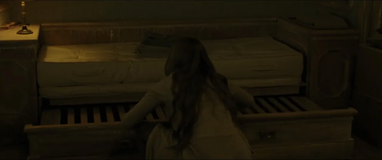 mother! (2017) movie still: medium — A dark, sparsely furnished room contains a low wooden bed with a pulled-out trundle. A woman with long hair…