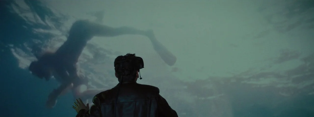 Dead Pigs (2018) movie still: medium — A man stands in a dark room facing a large, wall-sized projection of water. A blurred silhouette of a swimmer…