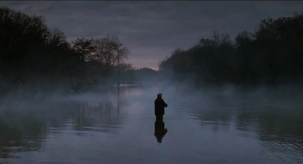Big Fish (2003) movie still: wide — A calm, misty river winds through a dark treeline during the early morning. An elderly man stands waist-deep in…