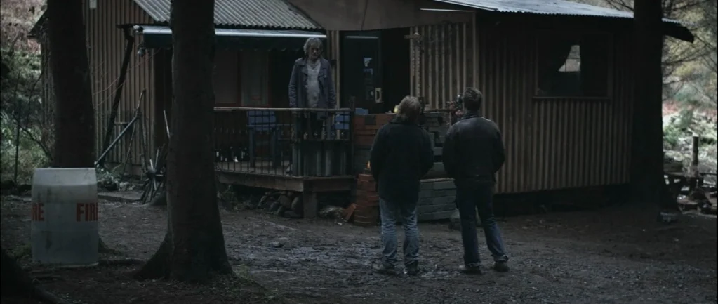 One Hundred Mornings (2009) movie still: wide — A wooden cabin stands in a dark forest clearing with a muddy ground surface. Three men interact in a…
