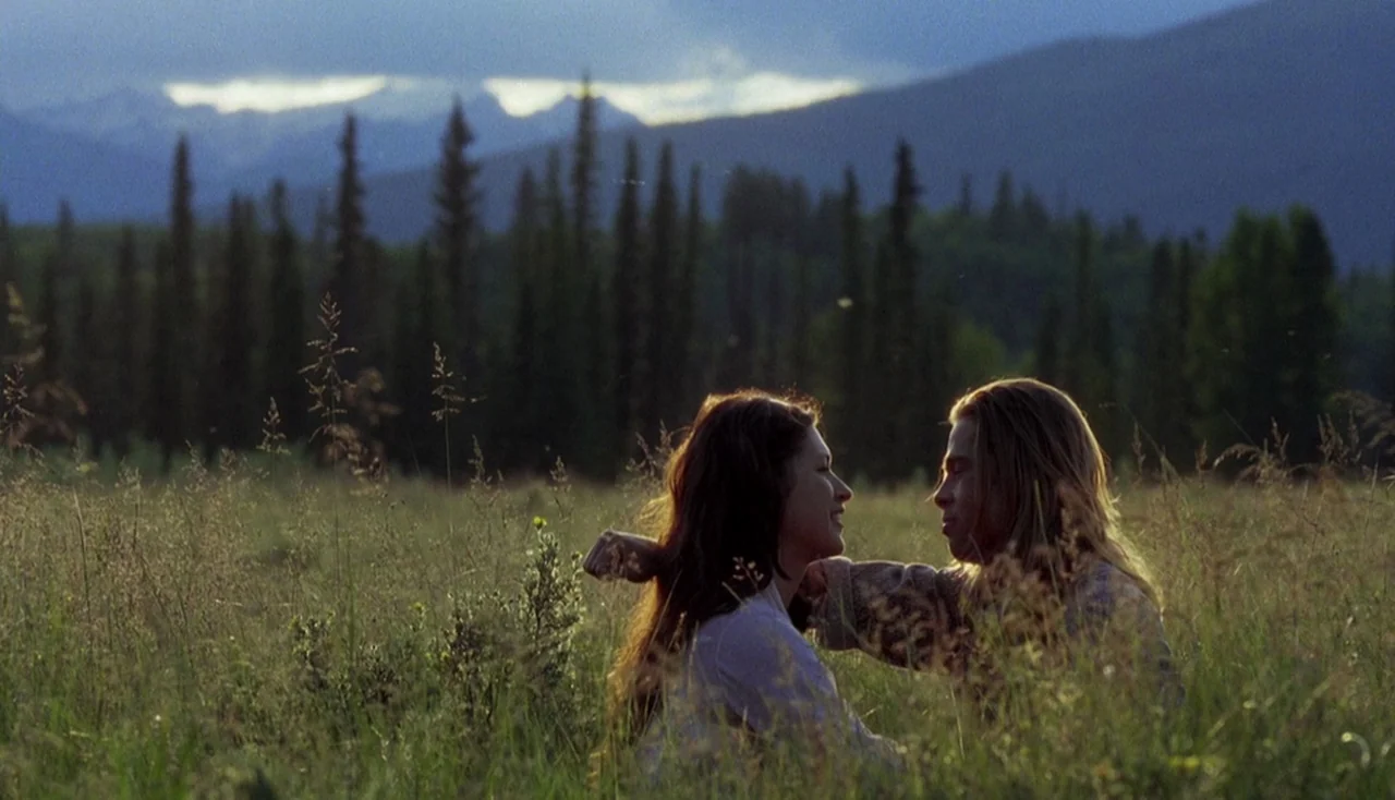 Legends of the Fall (1994) movie still: medium — A wide field of tall grass with coniferous trees and distant mountains in the back. A young man and…