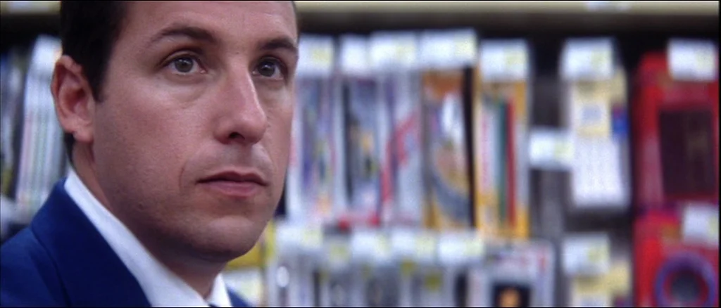 Punch Drunk Love (2002) movie still: closeup — A man stands in an aisle of a store with blurred retail shelves behind him. He gazes off-camera at an…