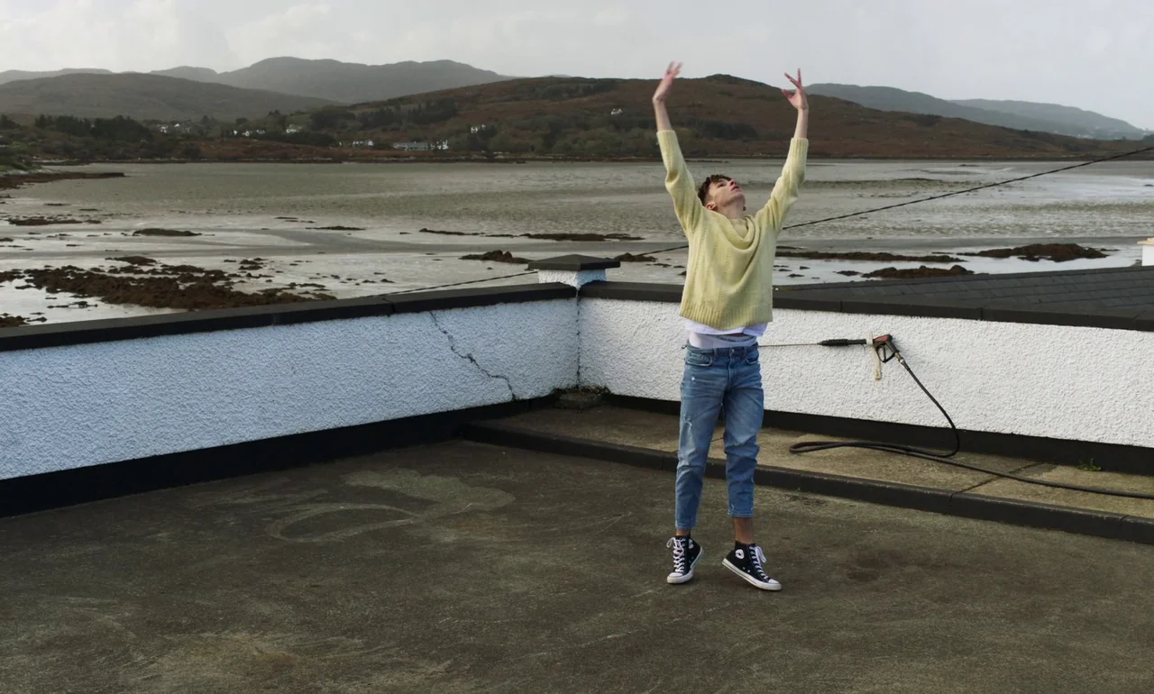 It Is In Us All (2022) movie still: fullbody — A flat concrete roof with a white parapet overlooks a large bay at low tide. A young man stands on…