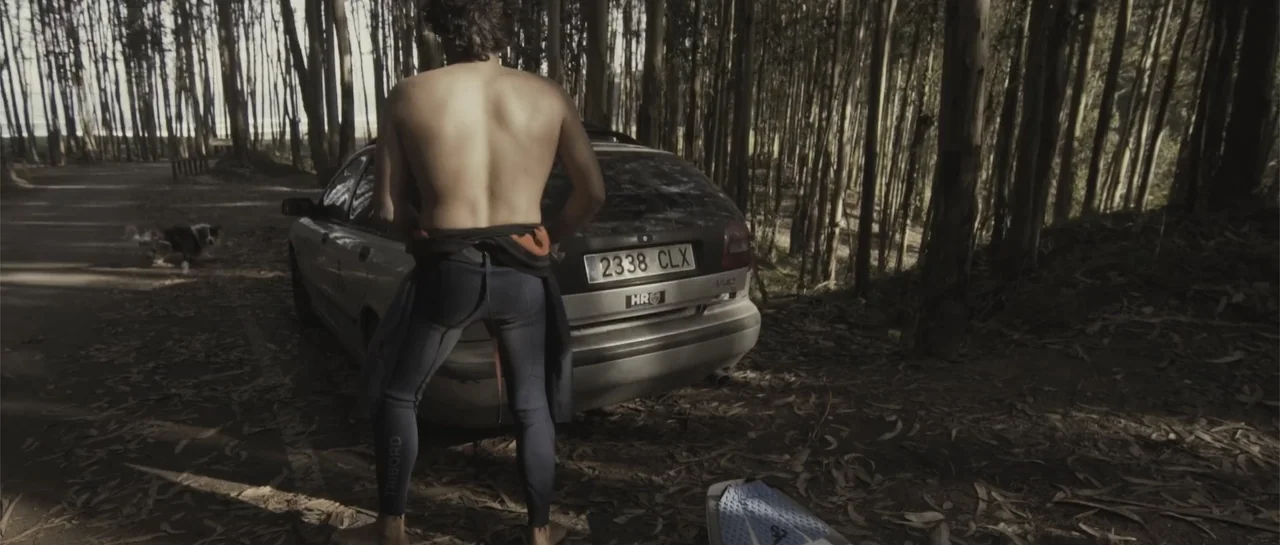 Taking Back the Waves (2017) movie still: fullbody — A dense grove of thin eucalyptus trees surrounds a silver station wagon parked on dirt. A…