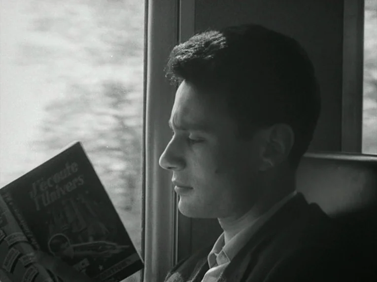 Le Petit Soldat (1963) movie still: closeup — A man sits by a train window inside a passenger car. He is looking down at a book titled J'écoute…