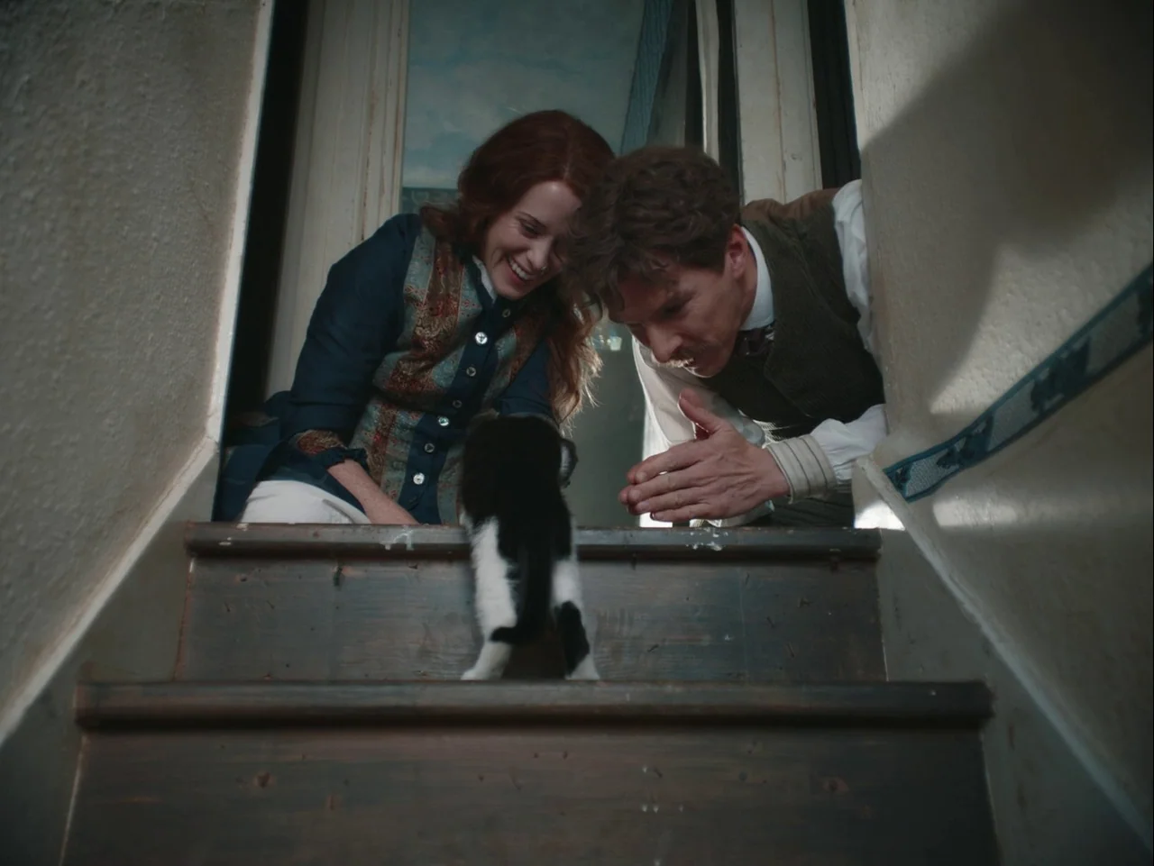 The Electrical Life of Louis Wain (2021) movie still: medium — A wooden staircase in a 1920s house with simple painted walls. A woman and man crouch…