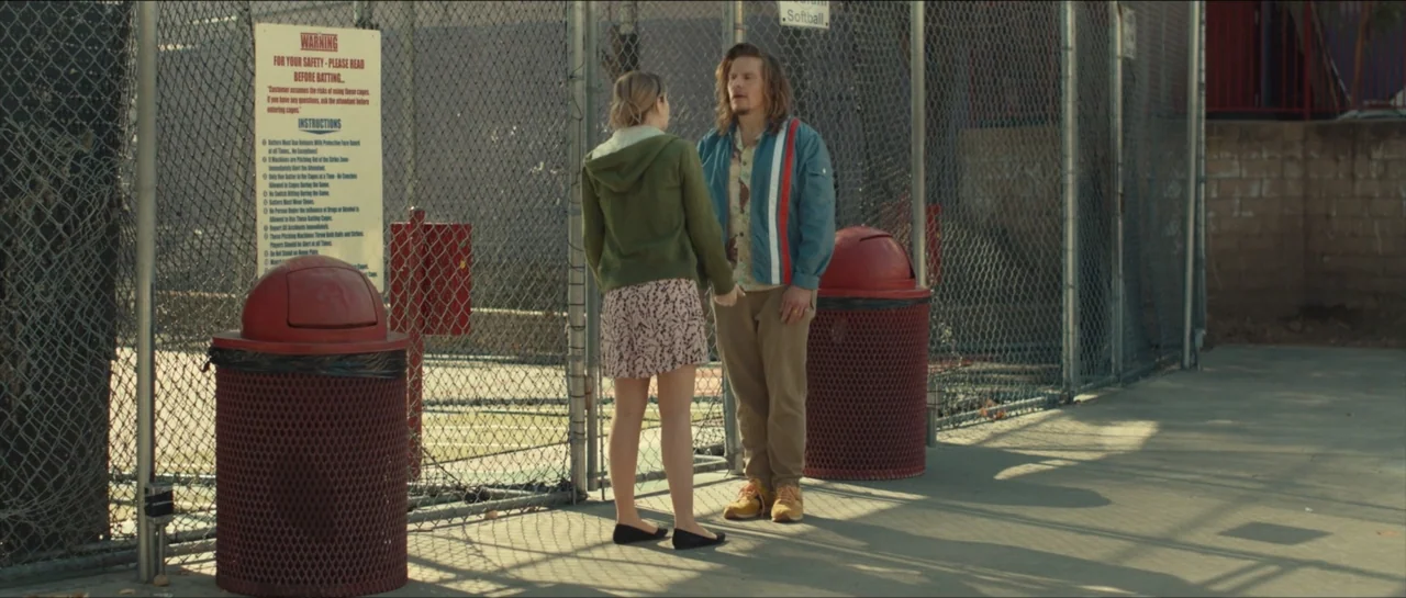 We Broke Up (2021) movie still: fullbody — A chain-link batting cage exterior with a prominent safety warning sign on the mesh. A man and a woman…
