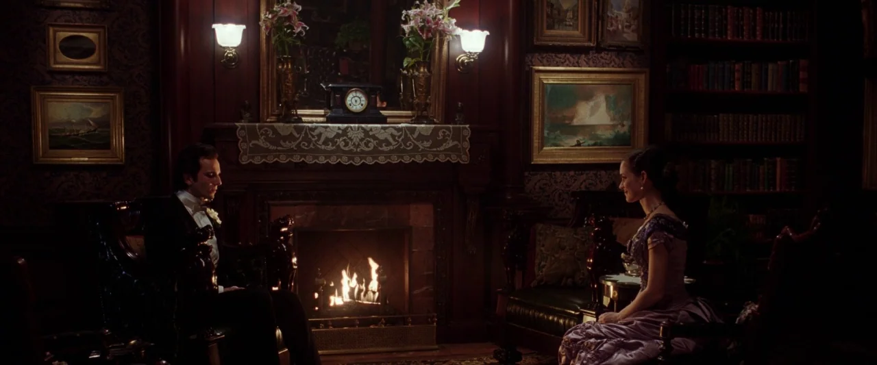 The Age of Innocence (1993) movie still: wide — A dark wood-paneled room features a central fireplace flanked by two plush armchairs. A man in…
