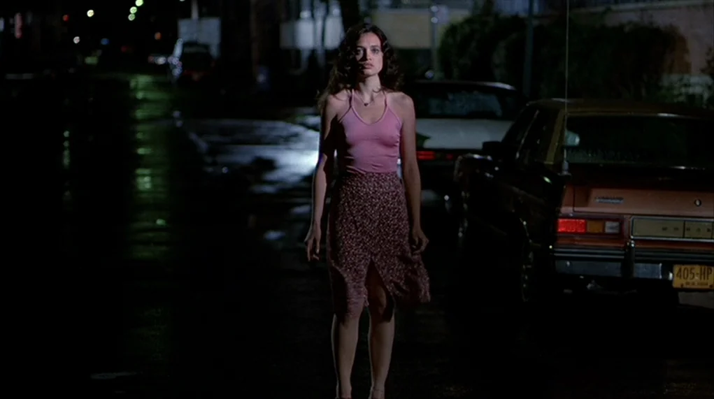 The Warriors (1979) movie still: fullbody — A nighttime city street features wet pavement reflecting light from distant lamps. A woman stands in the…