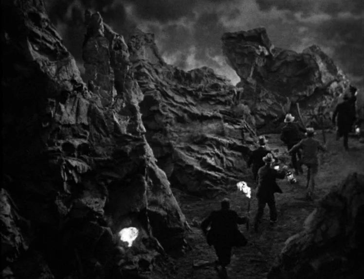Frankenstein movie still: wide — A dark, uneven expanse of jagged rock formations under a cloudy night sky. A group of men in suits runs through the…