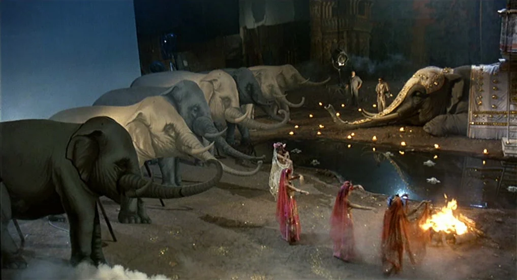 Intervista (1987) movie still: wide — A theatrical film set features multiple large artificial elephants positioned near a pool of water. Three…