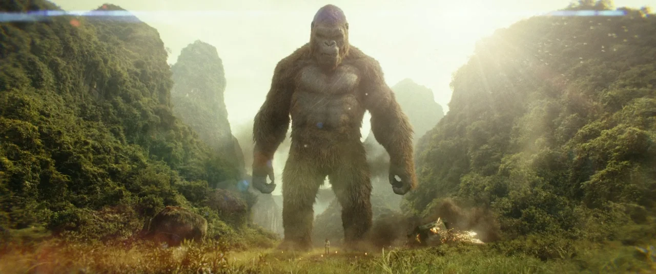 Kong: Skull Island (2017) movie still: wide — A massive gorilla stands in the center of a tropical valley between tall mountains. The creature looms…