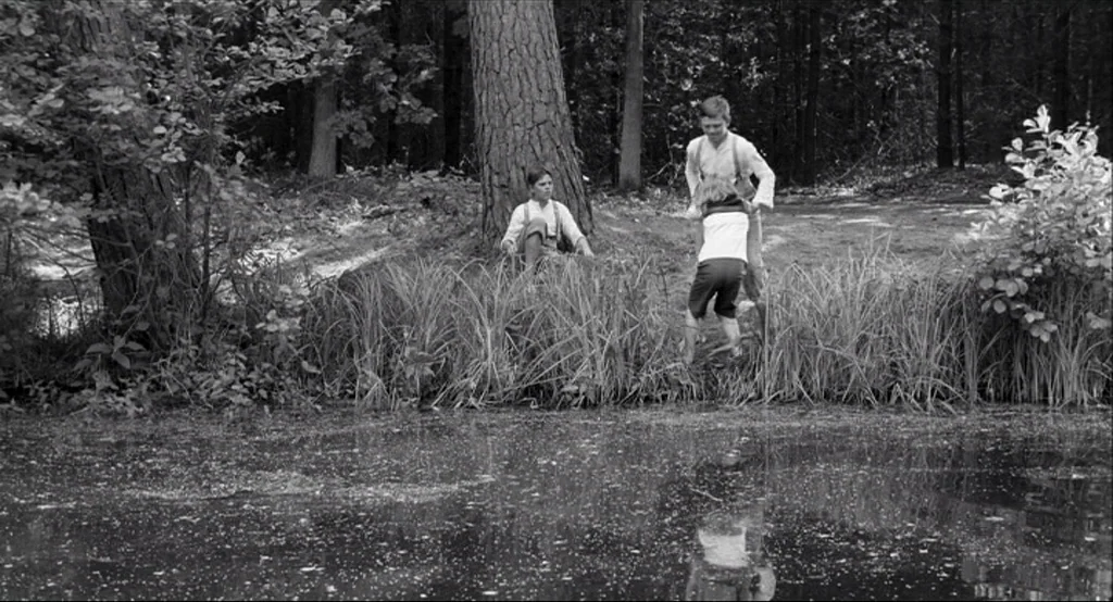 The White Ribbon (2009) movie still: wide — A group of three children stands at the muddy edge of a pond. One child leaps while the other two…