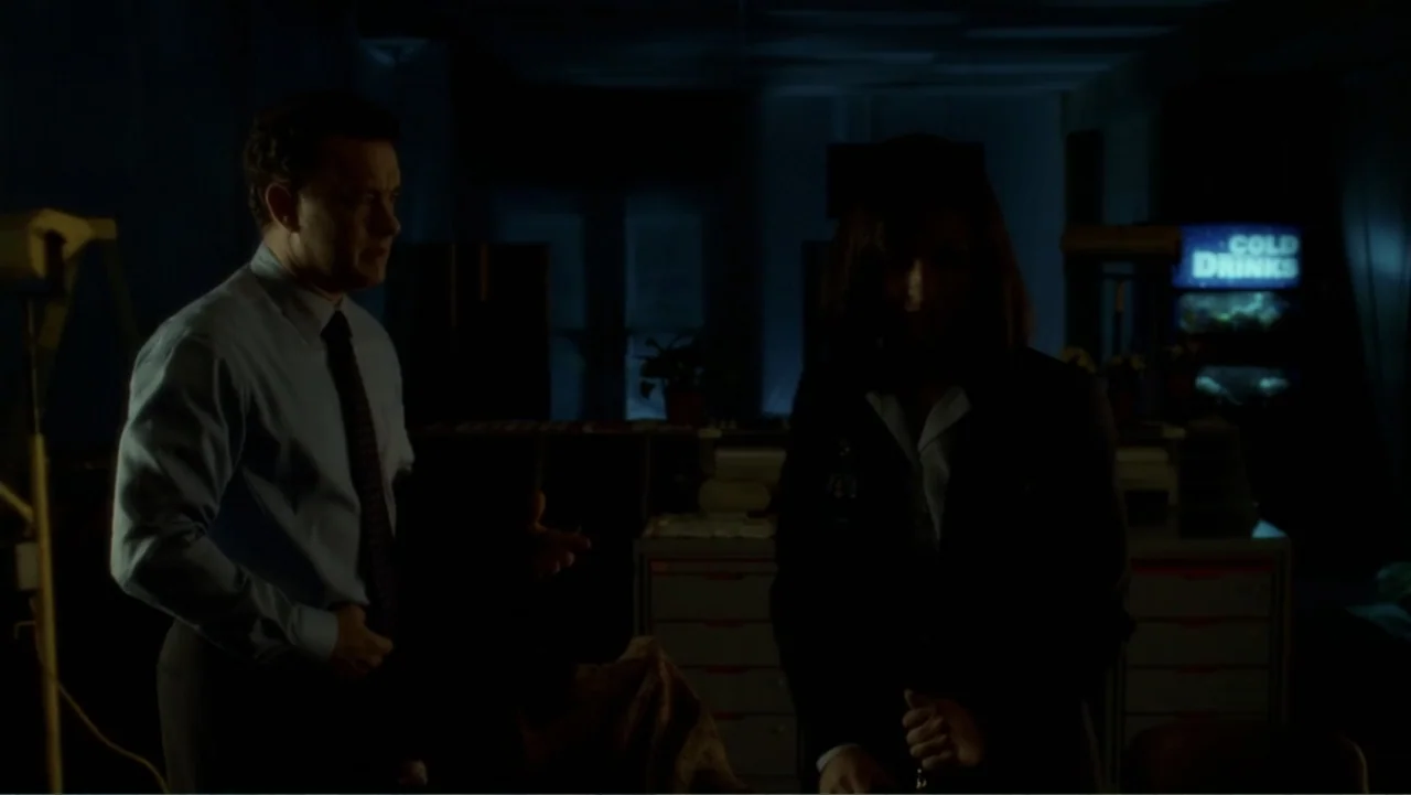 The Terminal (2004) movie still: medium — An interior office space is dimly lit at night with minimal ambient visibility. A man stands talking to a…