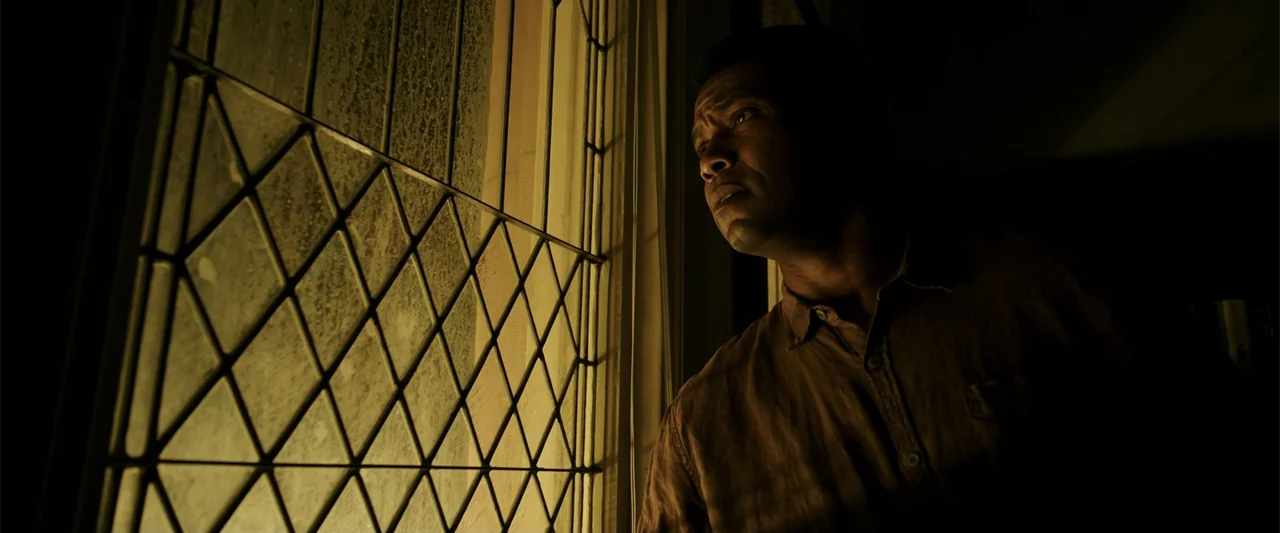K.G.F: Chapter 2 (2022) movie still: medium — A dimly lit room at night features a large window with a diamond-pattern metal lattice. A man stands…