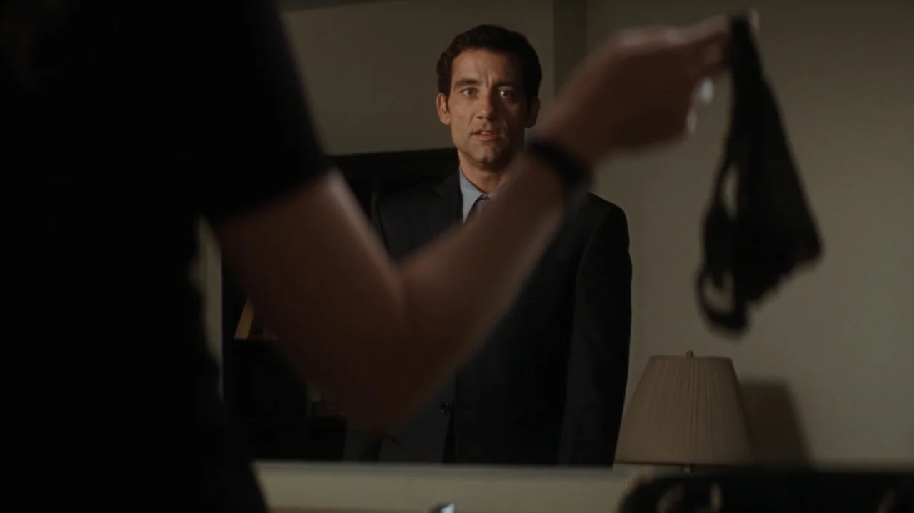 Duplicity (2009) movie still: medium — A modern room features a dark-haired man wearing a suit standing before a mirror. A woman in a black shirt…