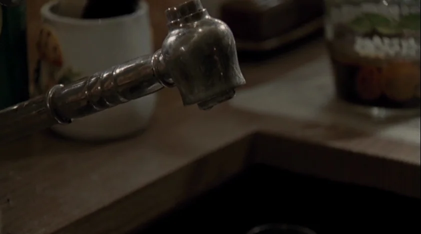 The House Of The Devil (2009) movie still: closeup — A close-up view of a metal kitchen faucet positioned above a dark basin. Decorative items sit…