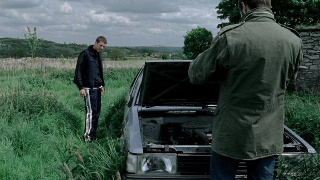 Dead Man’s Shoes (2004) movie still: medium — A silver hatchback sits with its hood open in a dense, green grassy field. A young man watches another…