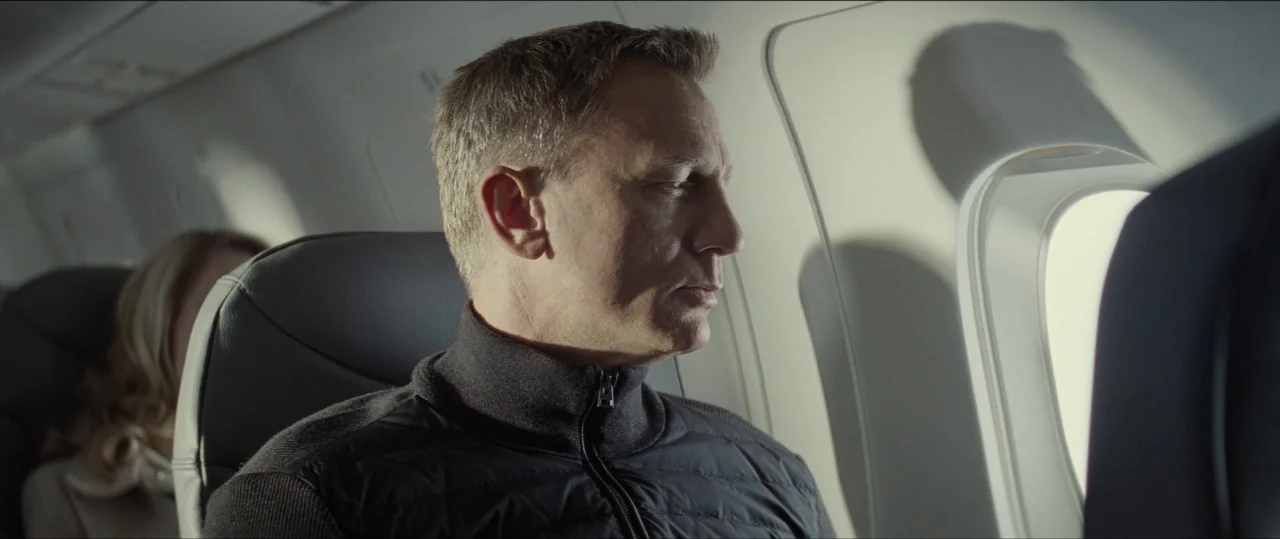 Spectre (2015) movie still: medium — A man sits in an airplane seat next to a bright window. He gazes outside at the view through the small oval…