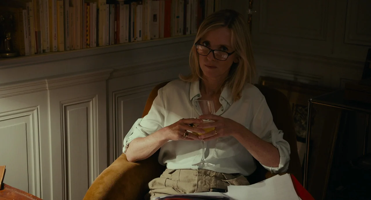 Last Summer (2023) movie still: medium — A dark study features a bookshelf filled with books and white wall paneling. A middle-aged woman with…