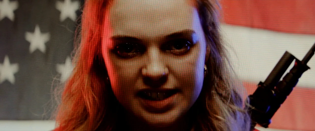 Assassination Nation (2018) movie still: closeup — A woman stands before a large American flag mounted on a wall. She stares intensely while lit by…