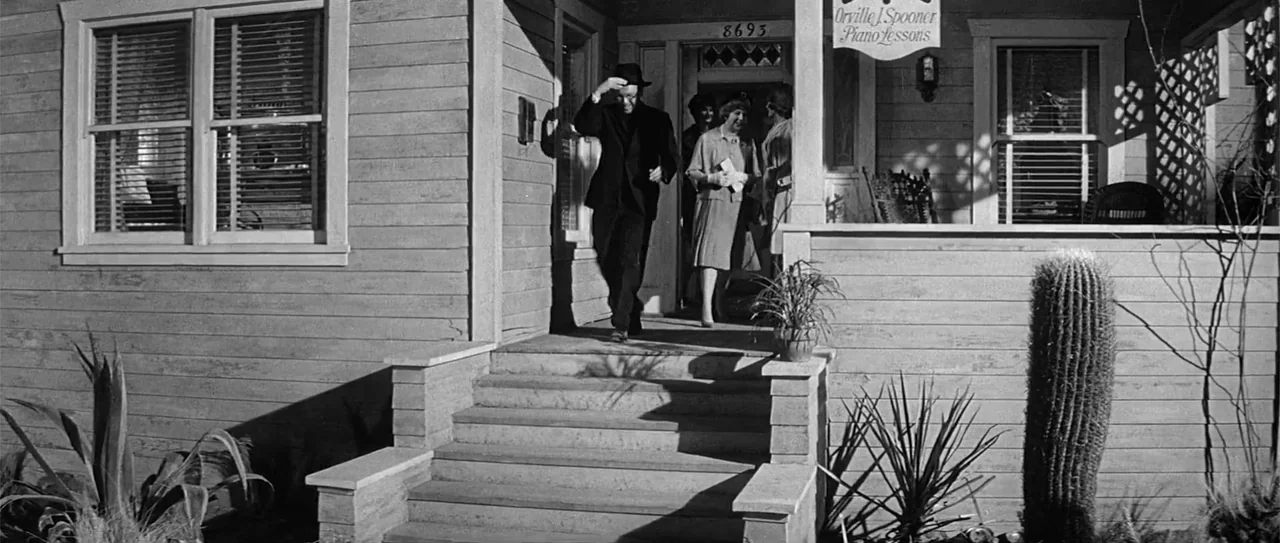 Kiss Me, Stupid (1964) movie still: fullbody — The wooden exterior of a suburban house features a porch with a cactus nearby. A man in a dark suit…