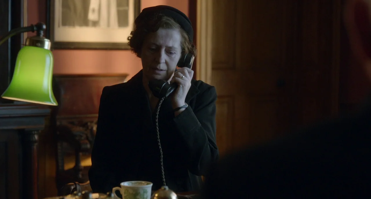 Brooklyn (2015) movie still: medium — A dimly lit room features a vintage desk with a green banker lamp. A woman sits in shadow holding a telephone…
