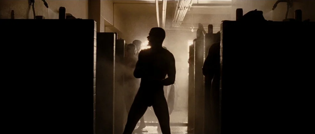 Jarhead movie still: fullbody — A long row of tiled shower stalls contains multiple silhouetted figures in a room. One man stands centrally in the…