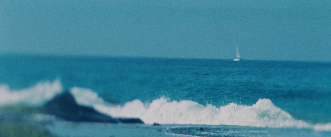 Savages (2012) movie still: establishing — A wide view of the ocean with waves breaking against dark foreground rocks. A small sailboat sits on the…
